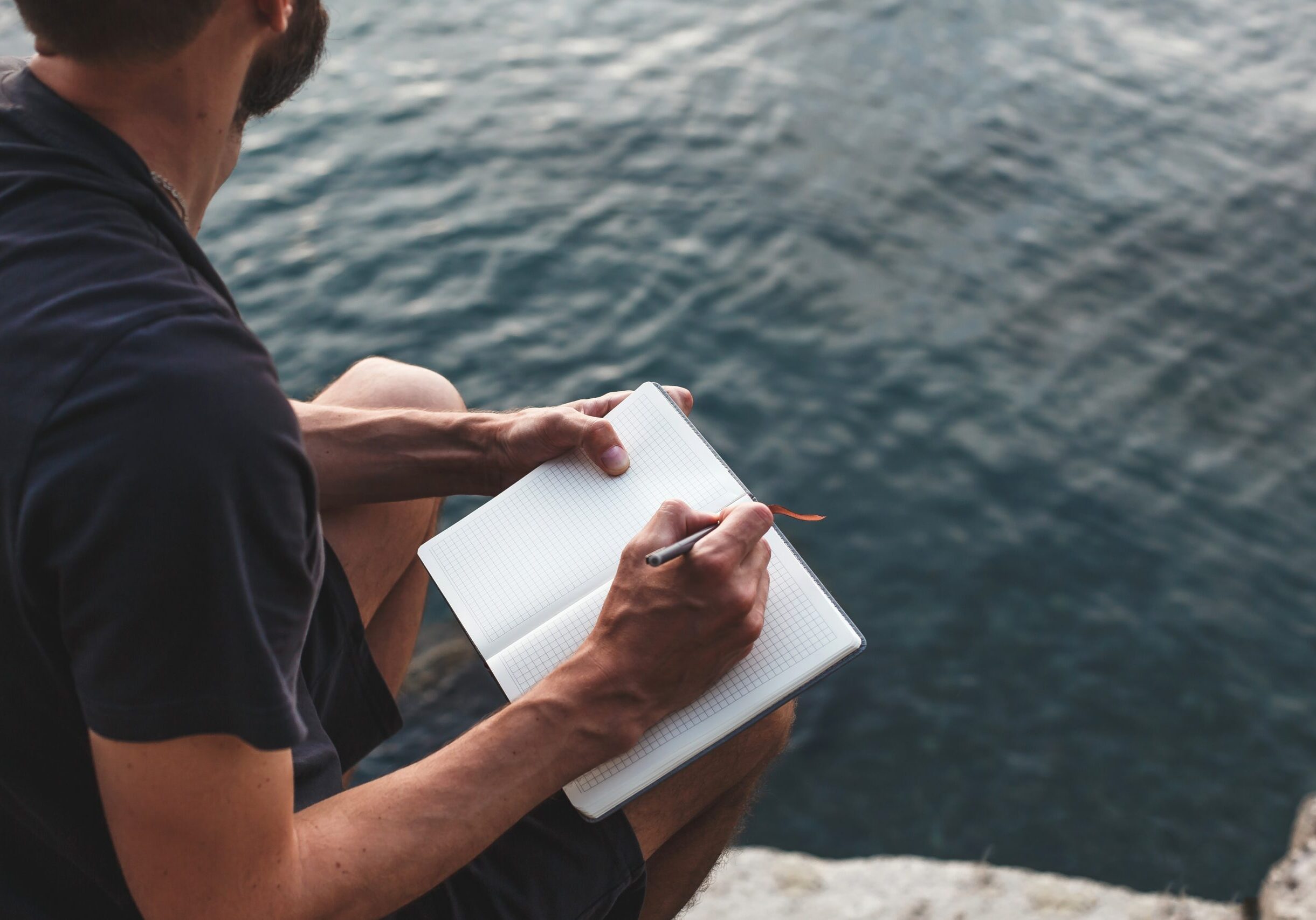 Man,Sits,On,A,Rocky,Shore,By,Water,,Writing,In